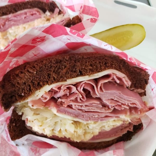 A deli sandwich with meat, cheese, and sauerkraut on dark bread, wrapped in checkered paper, with a pickle.
