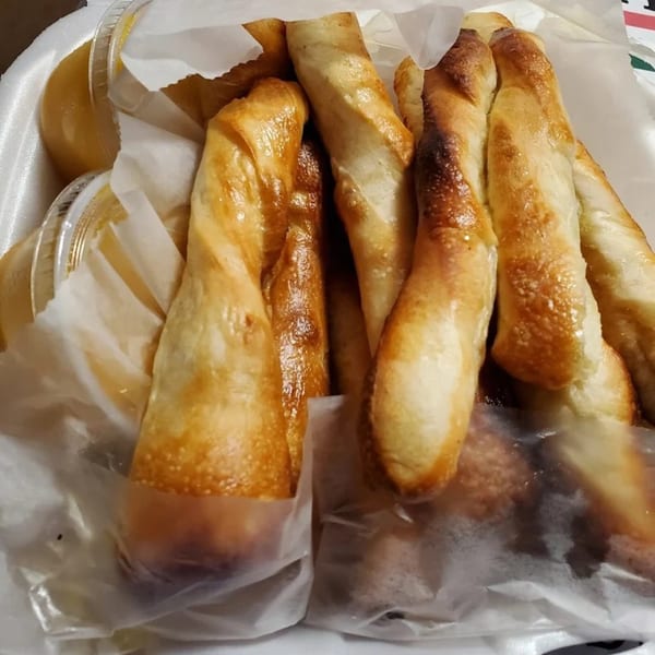 Golden-brown Catanzaro's breadsticks, some wrapped in paper, with dipping sauce in a white container.