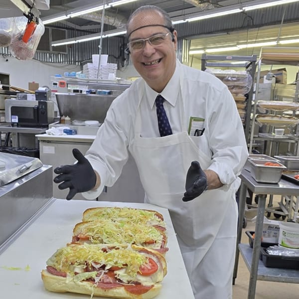 A smiling chef in a hairnet and apron presents three large, fully loaded subs in a commercial kitchen.