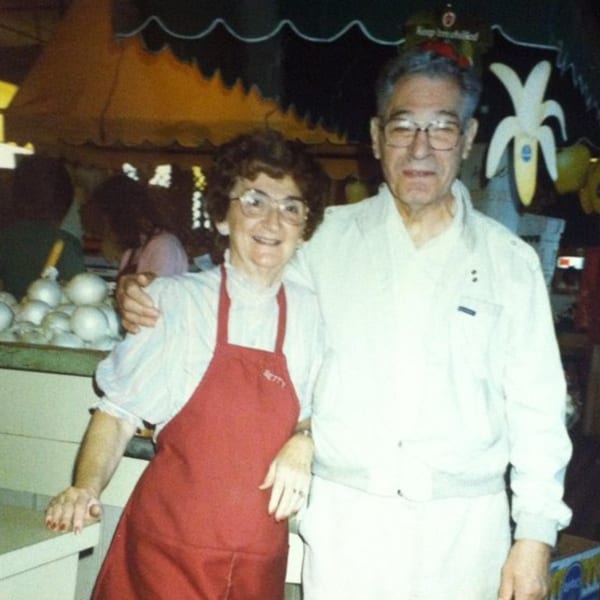 Betty, in a red apron, and a man smiling, with onions and a banana sign in the background.