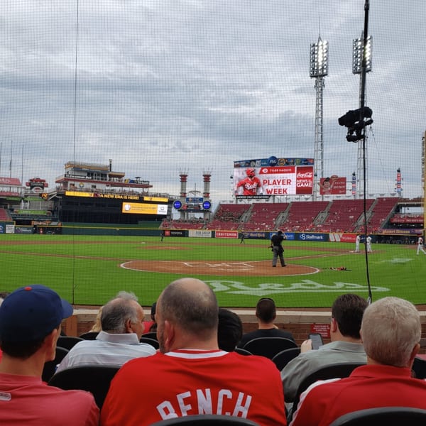 Spectators in red shirts watch a baseball game from behind a net, with stadium lights and screens visible.