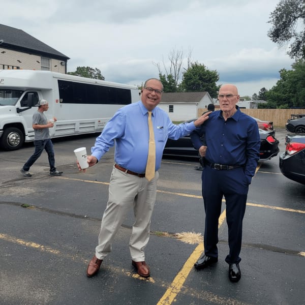 Two men in business casual attire stand in a parking lot, one with his arm around the other.